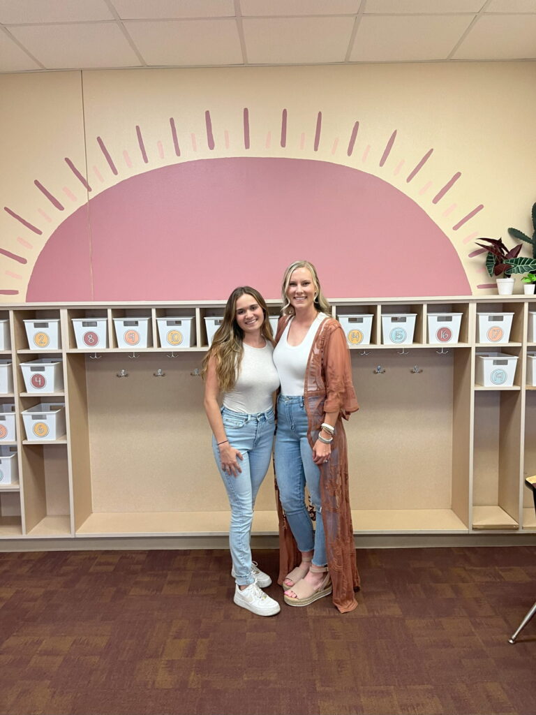 Shows a picture of Megan and Ally in front of the painted wall that has a sunset on it with a neutral color palette