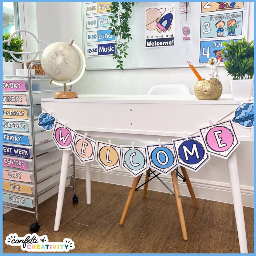 Shows an example of the space welcome banner on a white desk. The banner is held together by twine and clothespins, and the rest of the space classroom decor is in the background