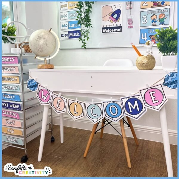 Space Welcome Banner 3 Shows an example of the space welcome banner on a white desk. The banner is held together by twine and clothespins, and the rest of the space classroom decor is in the background